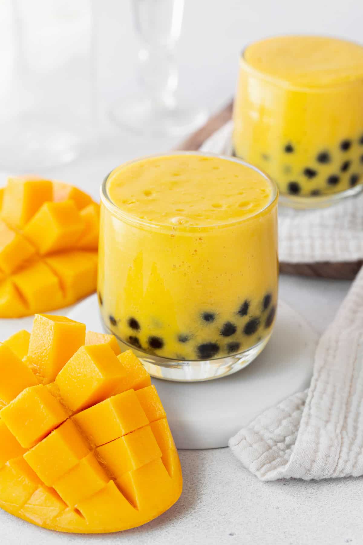 A closeup of a small glass of mango boba tea next to a cubed mango and towel.