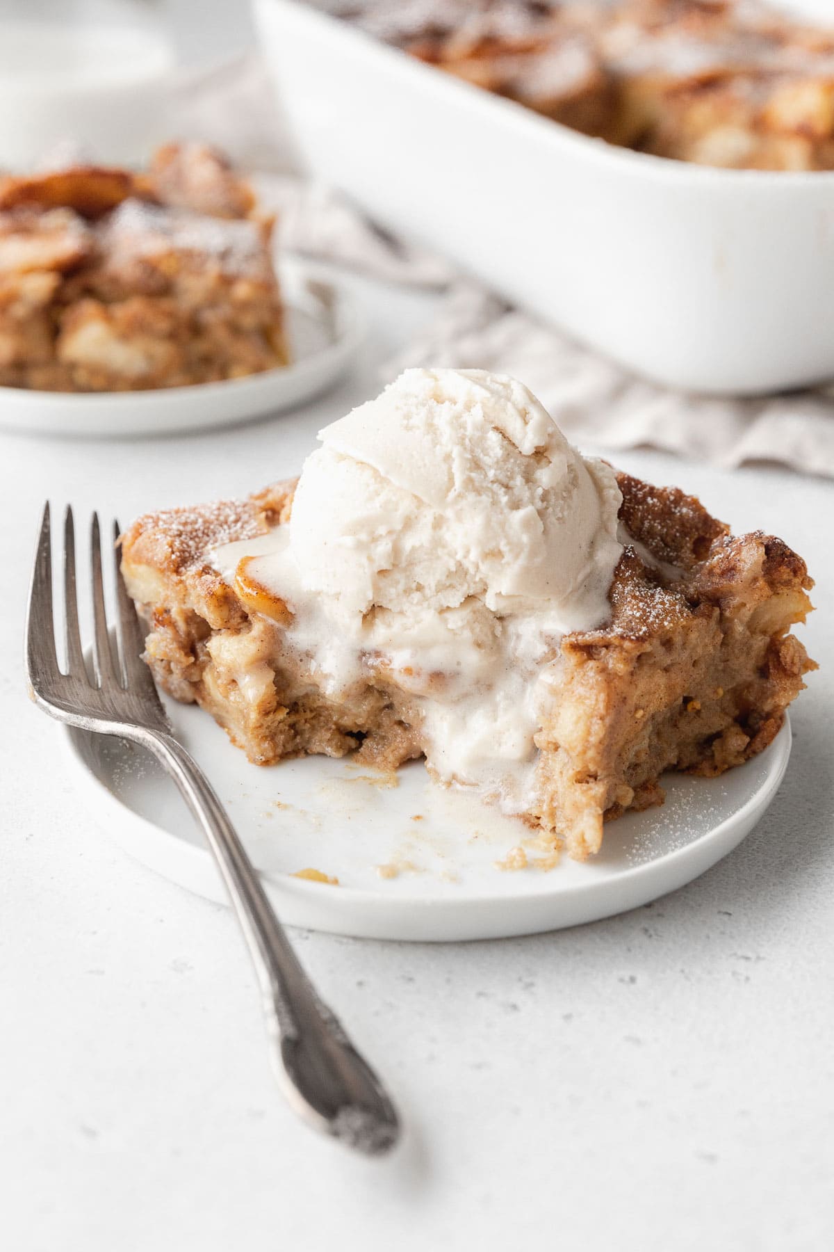 A partially eaten slice of vegan bread pudding on a white plate with a scoop of vanilla ice cream melting on top.