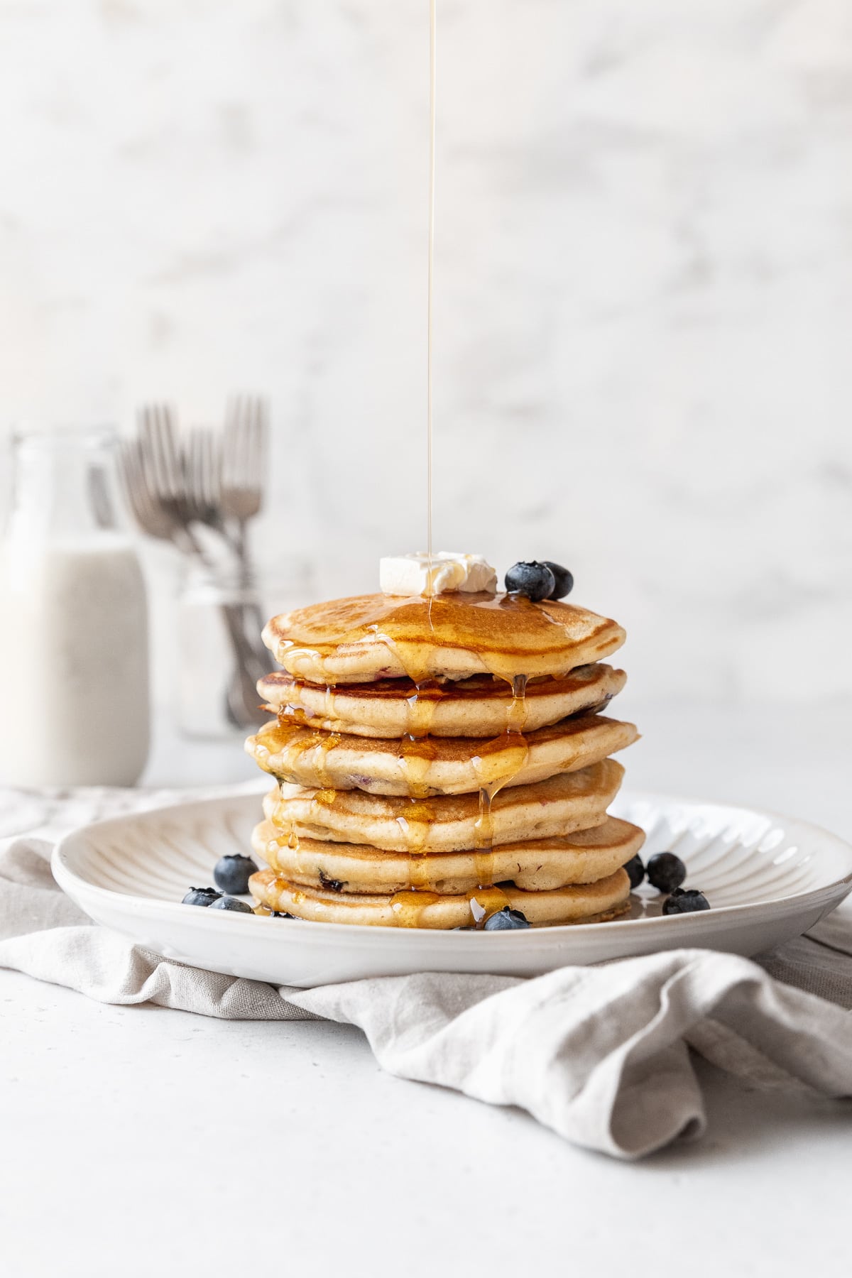 Distance side-on action shot of syrup being drizzled on a shortstack of blueberry studded muffin mix pancakes.