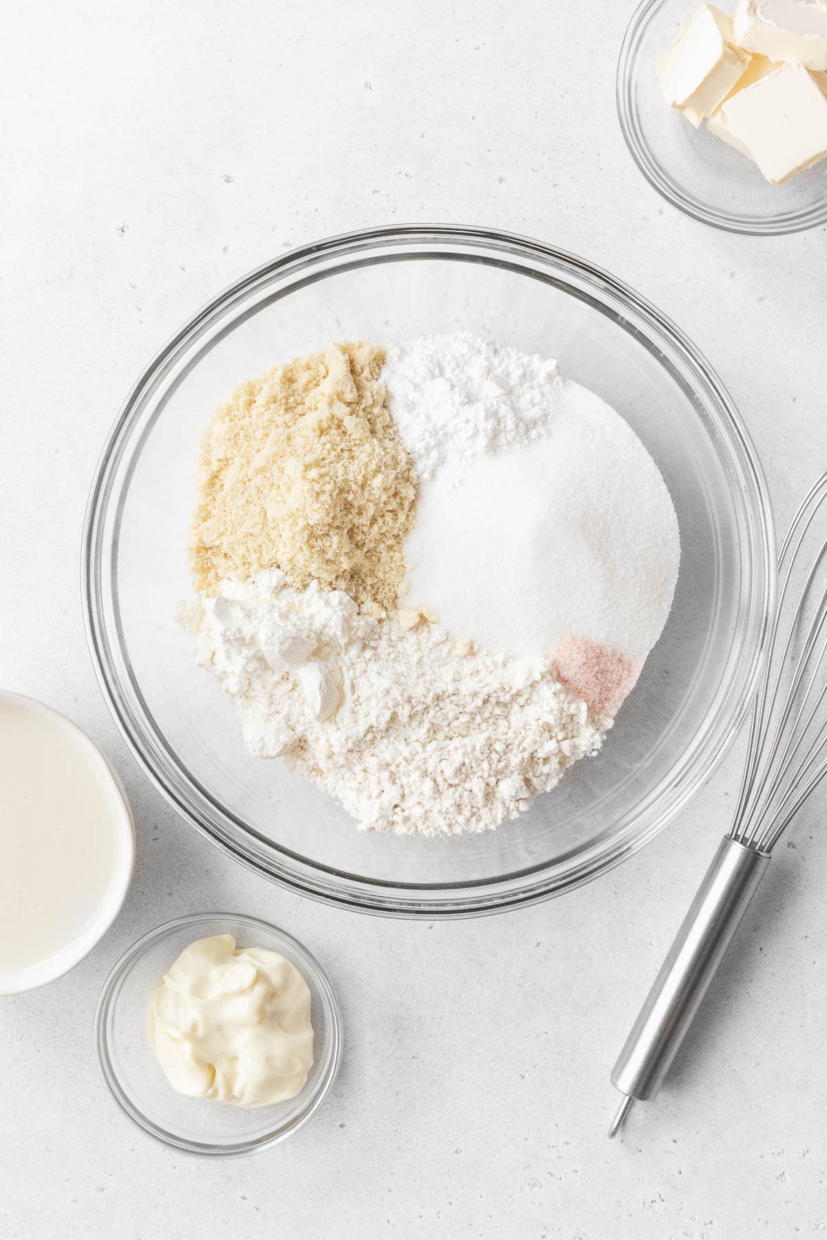 All of the dry ingredients for gluten-free and dairy-free tres leches cake in a glass mixing bowl with a whisk.  The other cake ingredients sit beside the mixing bowl.