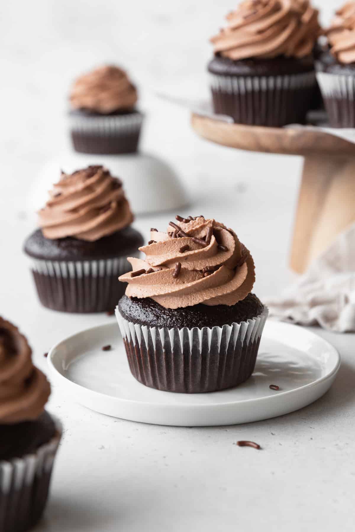 A chocolate cupcake with chocolate frosting on a white plate, with more cupcakes scattered in the background.