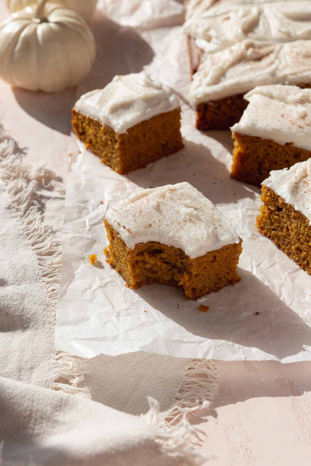 A bitten square of pumpkin bar with cream cheese frosting  on a piece of parchment paper with more squares scattered behind.
